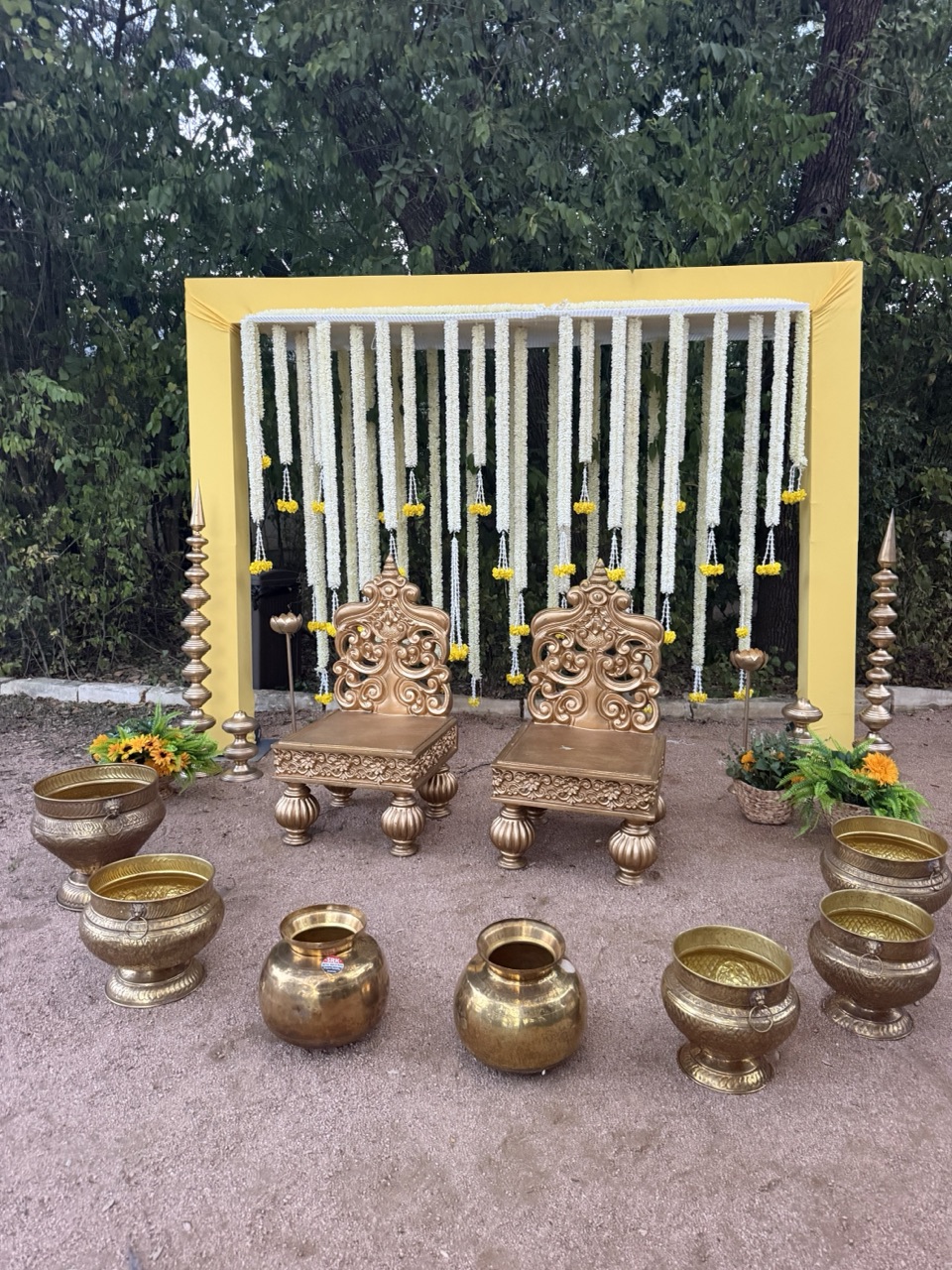 An outdoor frame of jasmine and marigold strands with carved gold chairs and brass kalash pots