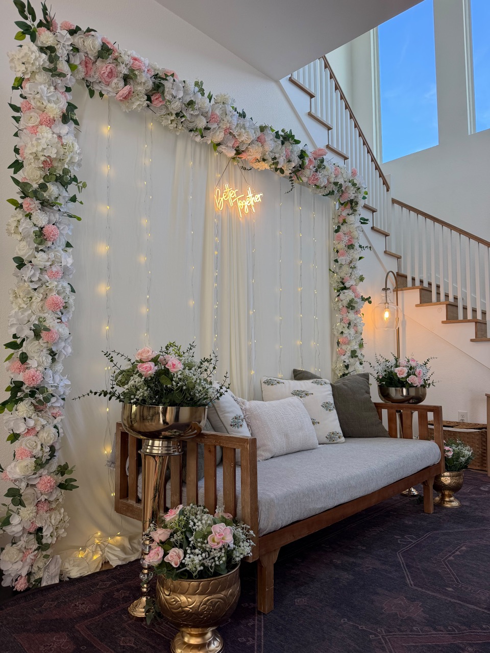 A home engagement setting with a floral arch, fairy light curtain and a neon sign reading Better Together