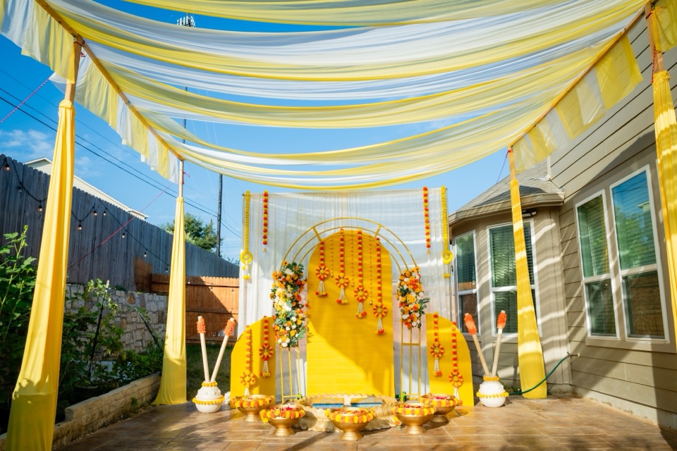 A backyard haldi under yellow and white canopy drapes against a blue sky, with a golden arch backdrop, marigold medallions and brass urlis