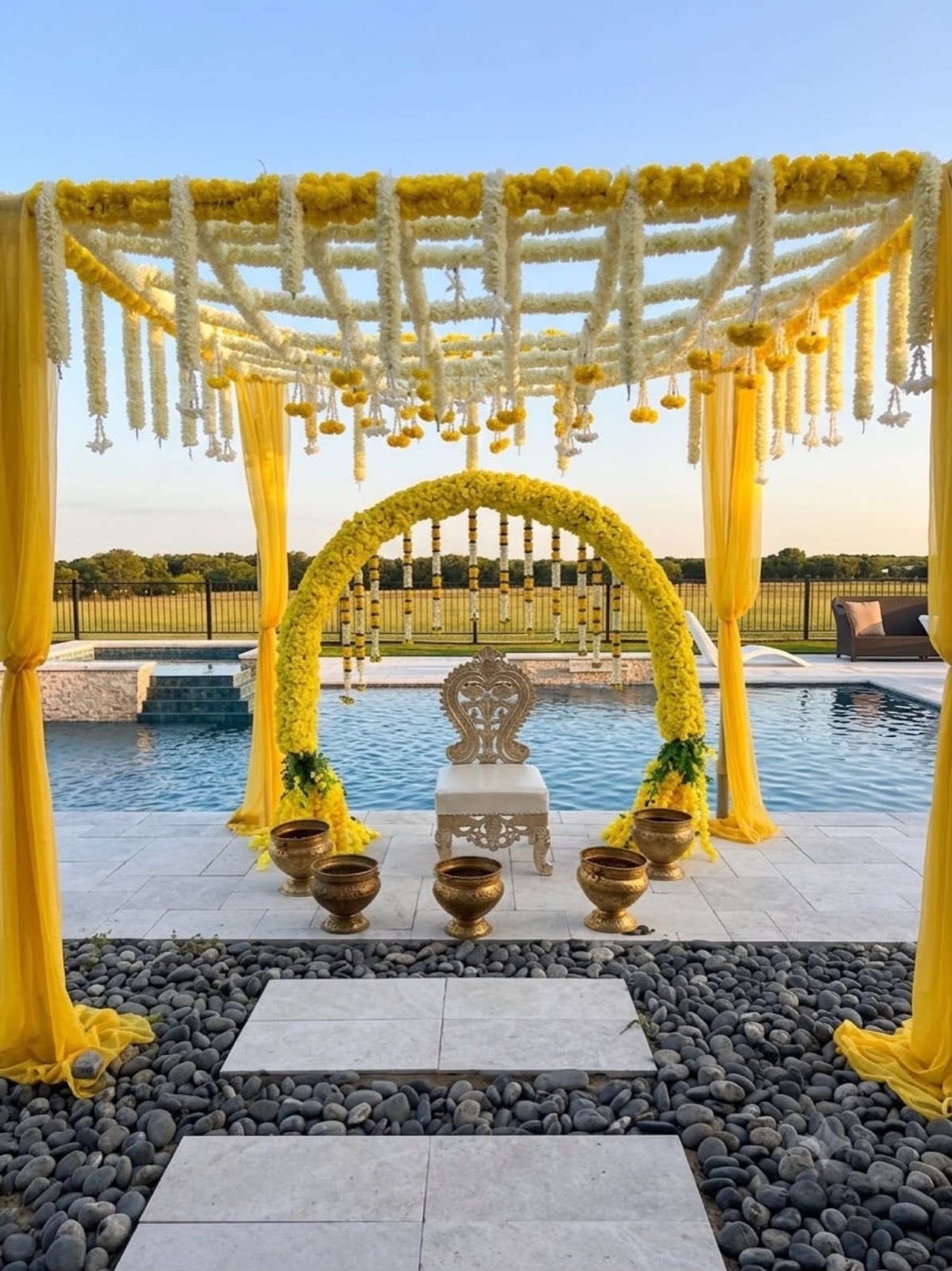 A poolside haldi at sunset with a marigold and jasmine lattice canopy, a yellow floral arch and brass urlis