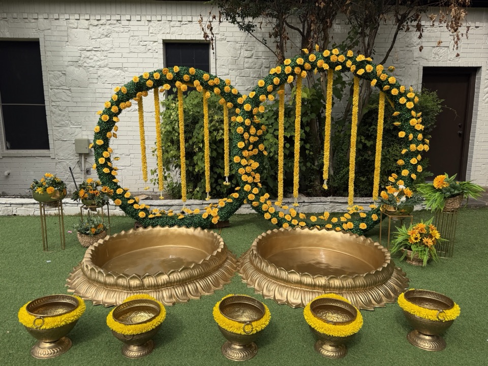 Two interlocking greenery rings with yellow roses and garlands above gold lotus urlis for a haldi