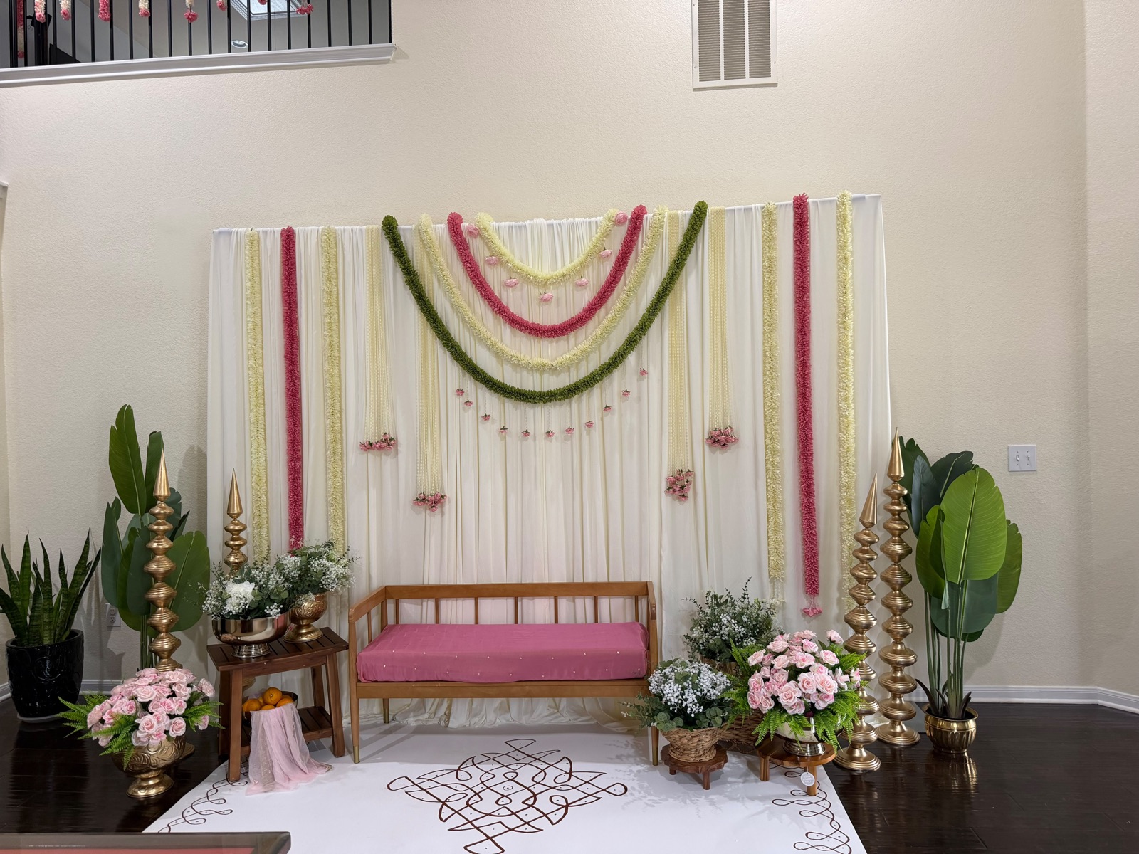 An ivory draped home ceremony backdrop with rose and green garland arcs above a hand drawn kolam