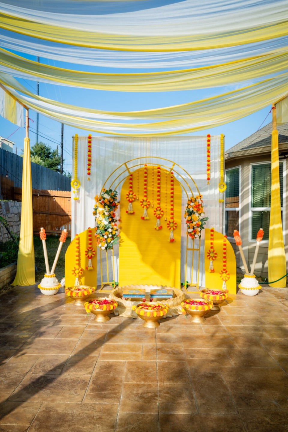 Backyard haldi under yellow and white canopy drapes, with a golden arch backdrop and marigold medallions