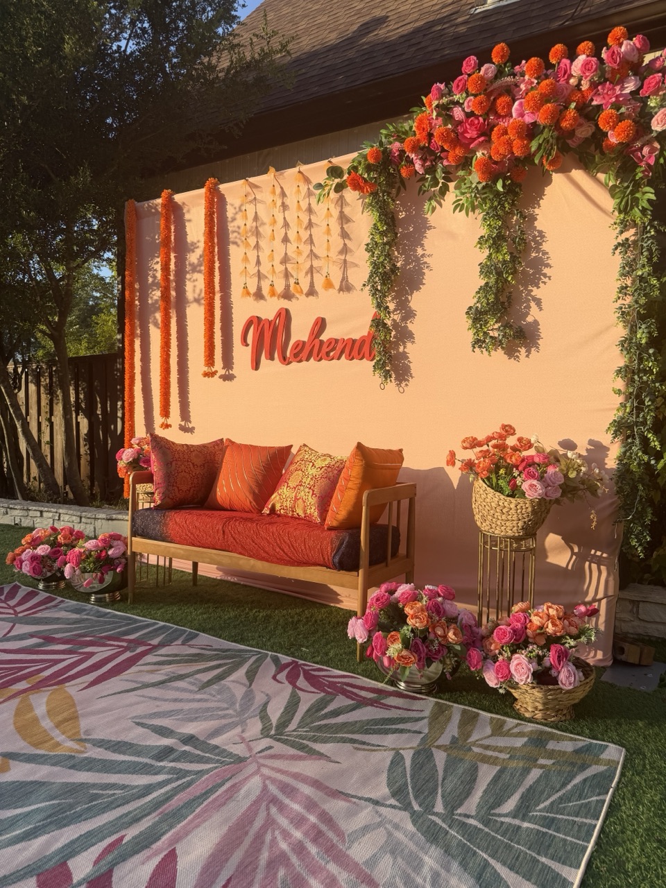 A golden hour mehendi with a peach backdrop, marigold strands, orange cushioned daybed and pink floral corners