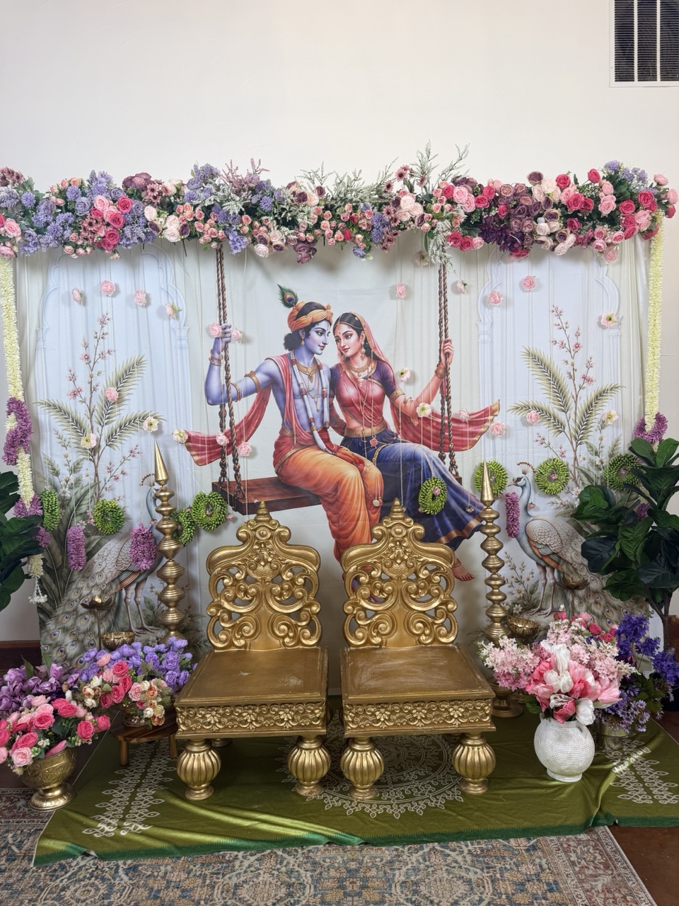 A Radha Krishna swing backdrop with purple and pink florals and carved gold seats