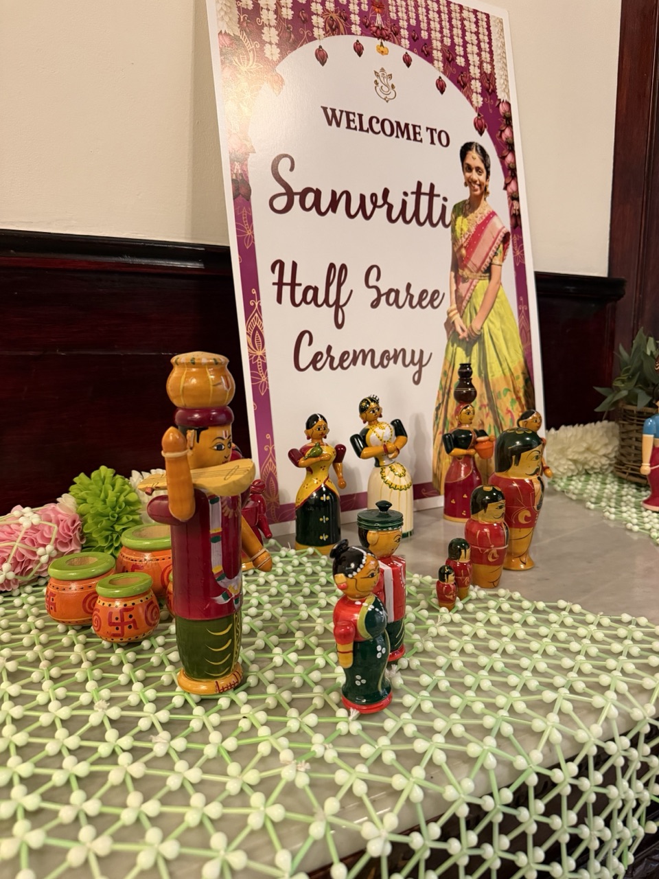 A half saree ceremony welcome board surrounded by traditional painted wooden dolls on a jasmine lattice runner